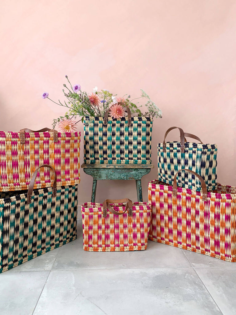 Group of handwoven chequered reed basket in pink, orange and green in front of a pink wall. One basket filled with flowers.