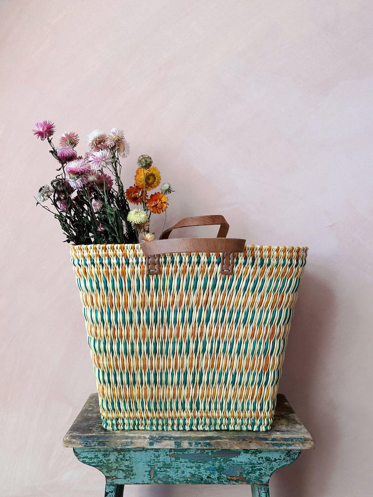 A tall rectangular shopper basket bag for wholesale, woven from natural reeds by Moroccan artisans featuring leather handles.