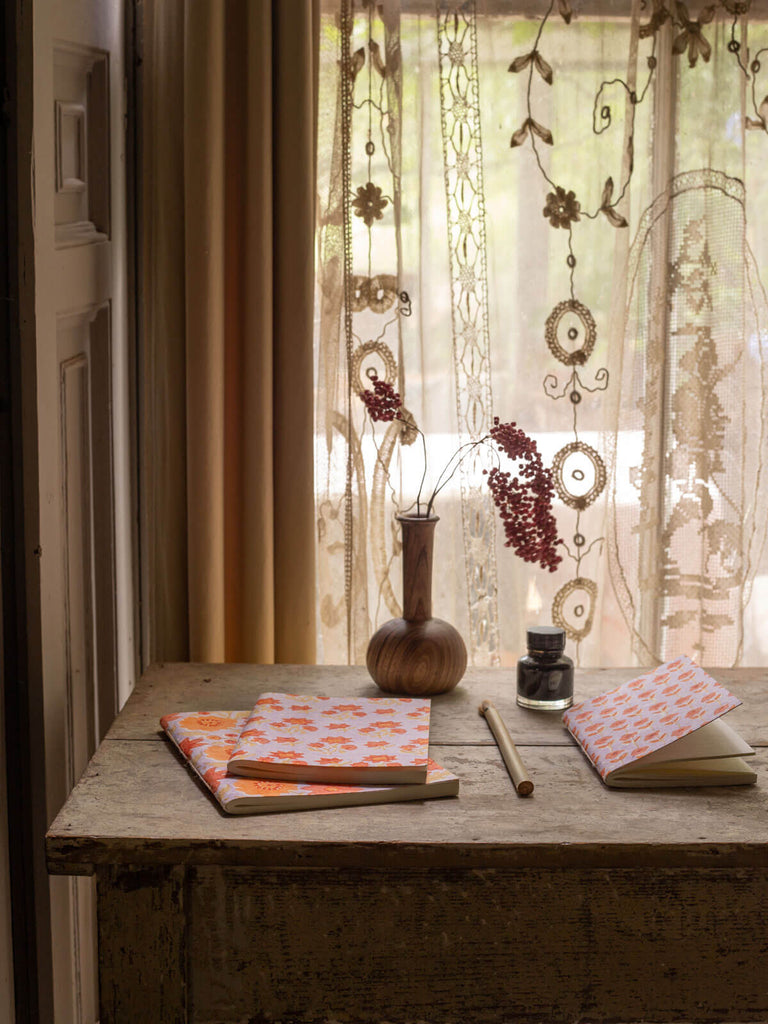 A trio of cotton paper notebooks with lilac and orange floral block printed covers on an antique desk
