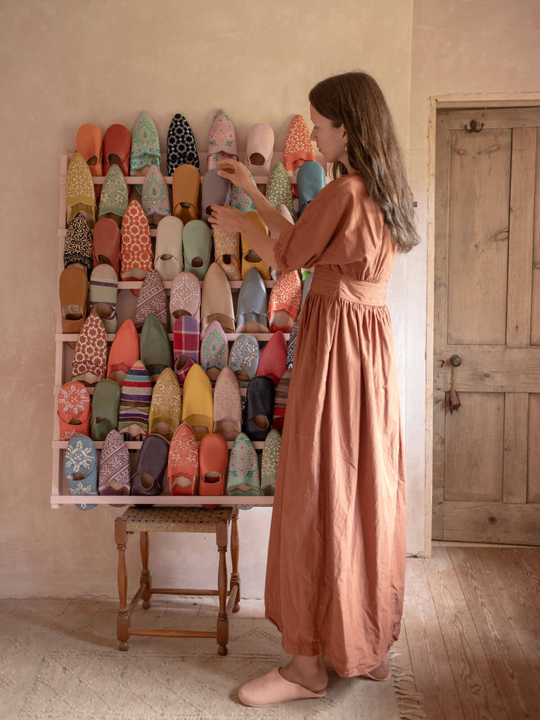 Woman in peach dress and pair of babouche slippers setting up a display of many different Bohemia Moroccan babouche slippers in a rustic plaster pink room.