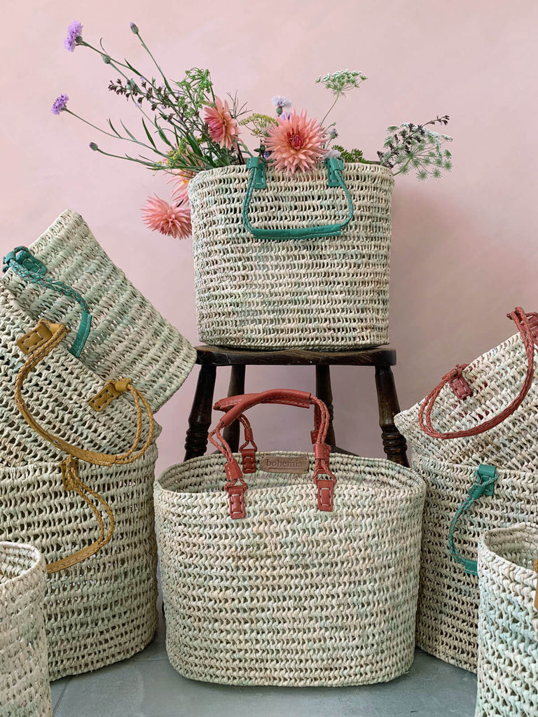 Oval shaped baskets with pleated leather handles in different colours filled with flowers in front of a pink wall