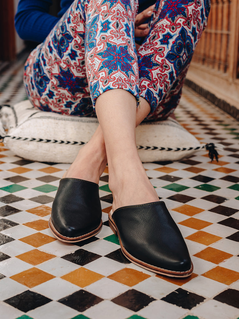 Woman wearing Leather mules in black leather by Bohemia Design with colourful trousers, sitting on Moroccan tiled floor