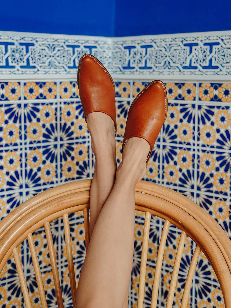 Woman wearing Leather tan mules by Bohemia Design while reclining on a cane chair against blue Moroccan tiles