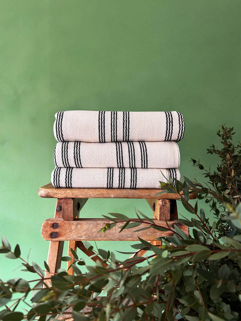 Stack of Ankara hammam towels, showcasing the soft cotton texture and a contrasting charcoal stripe, neatly folded on a wooden stool