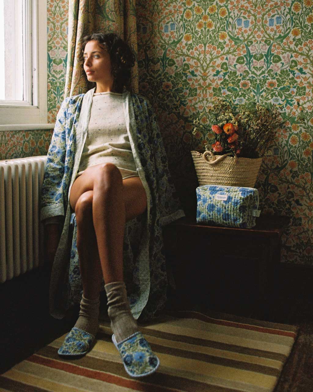 Model sitting at a window wearing a Bohemia block print cotton robe in sage and quilted house slippers in a country house