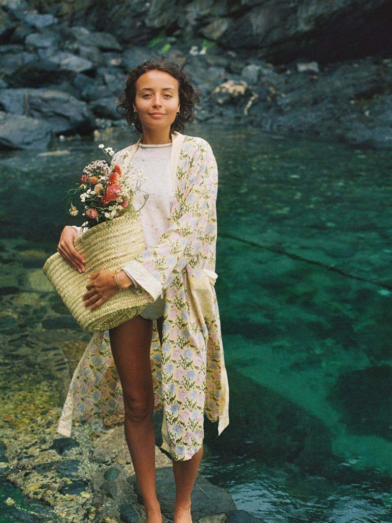 Model by the sea wearing a floral buttermilk block print robe holding a basket full of flowers.