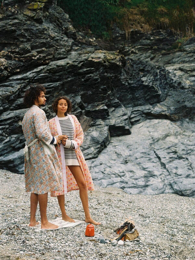 A male and female model on a pebble beach wearing duck egg and lilac block print robes by a campfire.