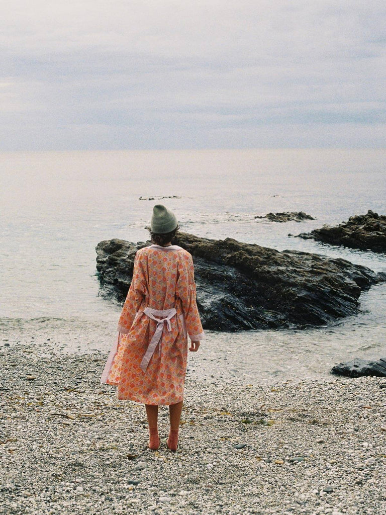 Model looking out to sea on a pebble beach wearing a floral lilac block print robe with cosy wool hat.