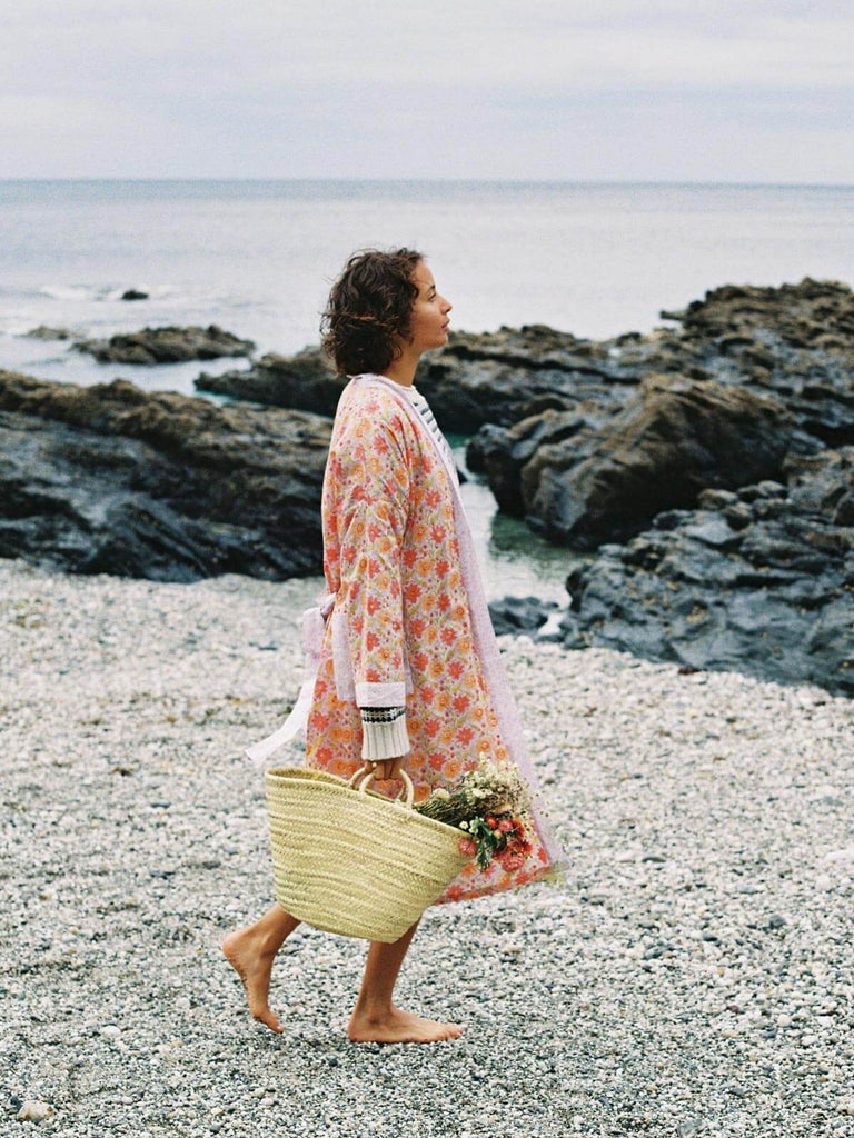 Model walking along a pebble beach wearing a lilac block print robe and Bohemia basket filled with dried flowers