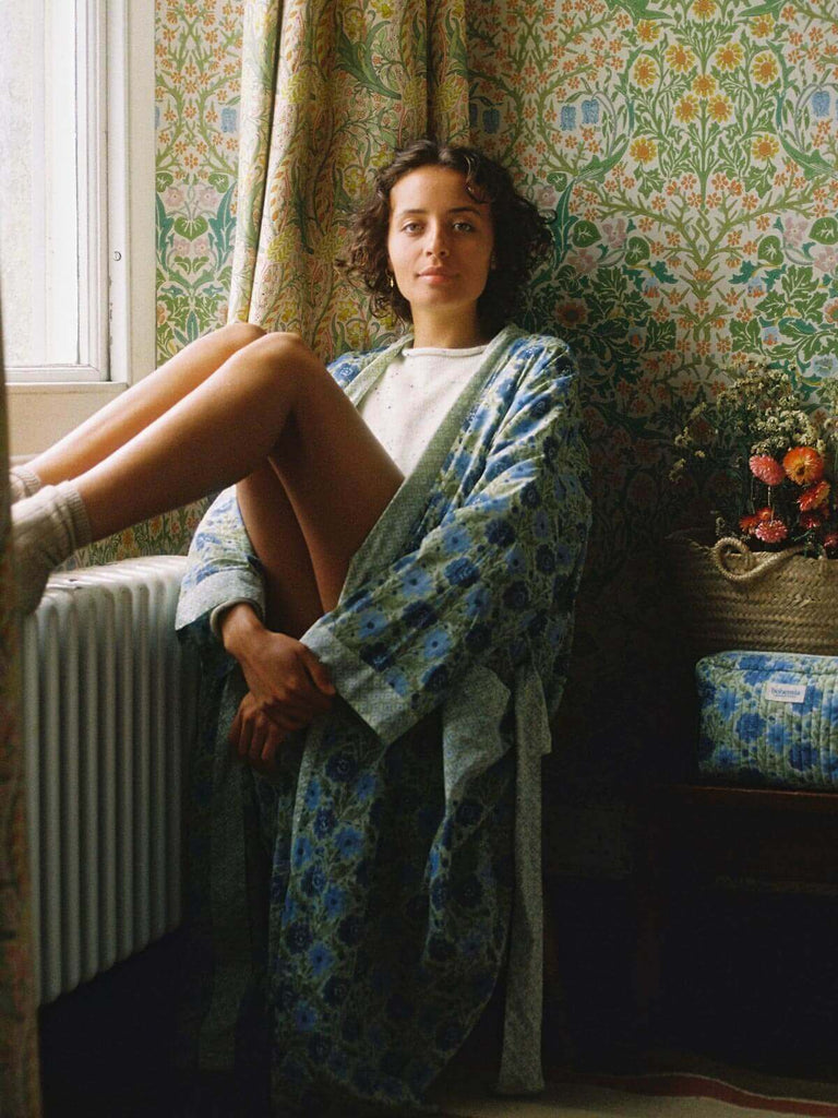 Model sitting at a window in a country house wearing sage block print robe and matching quilted house slippers