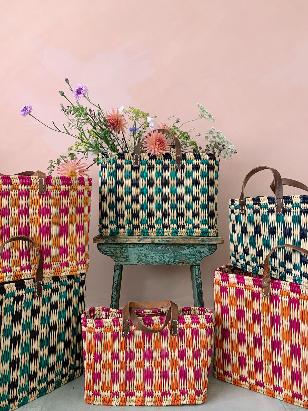 Group of handwoven chequered reed basket in pink, orange and green in front of a pink wall. One basket filled with flowers.