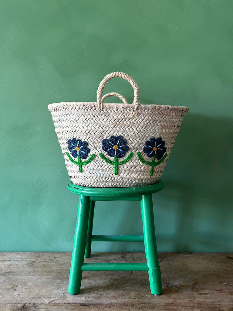 Handwoven basket with embroidered daisies, placed on a green stool against a vibrant green wall | Bohemia Design