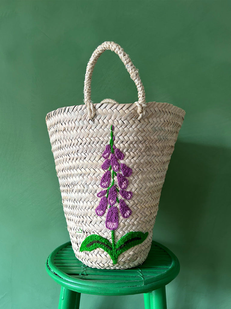 Natural woven basket bag with a hand-embroidered foxglove flower on a green stool against a green wall by Bohemia Design