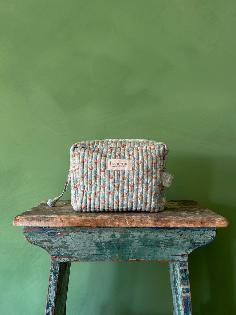 Bohemia Design washbag with a garland floral pattern in duck egg green, blue, and terracotta, resting on a rustic stool against a green wall