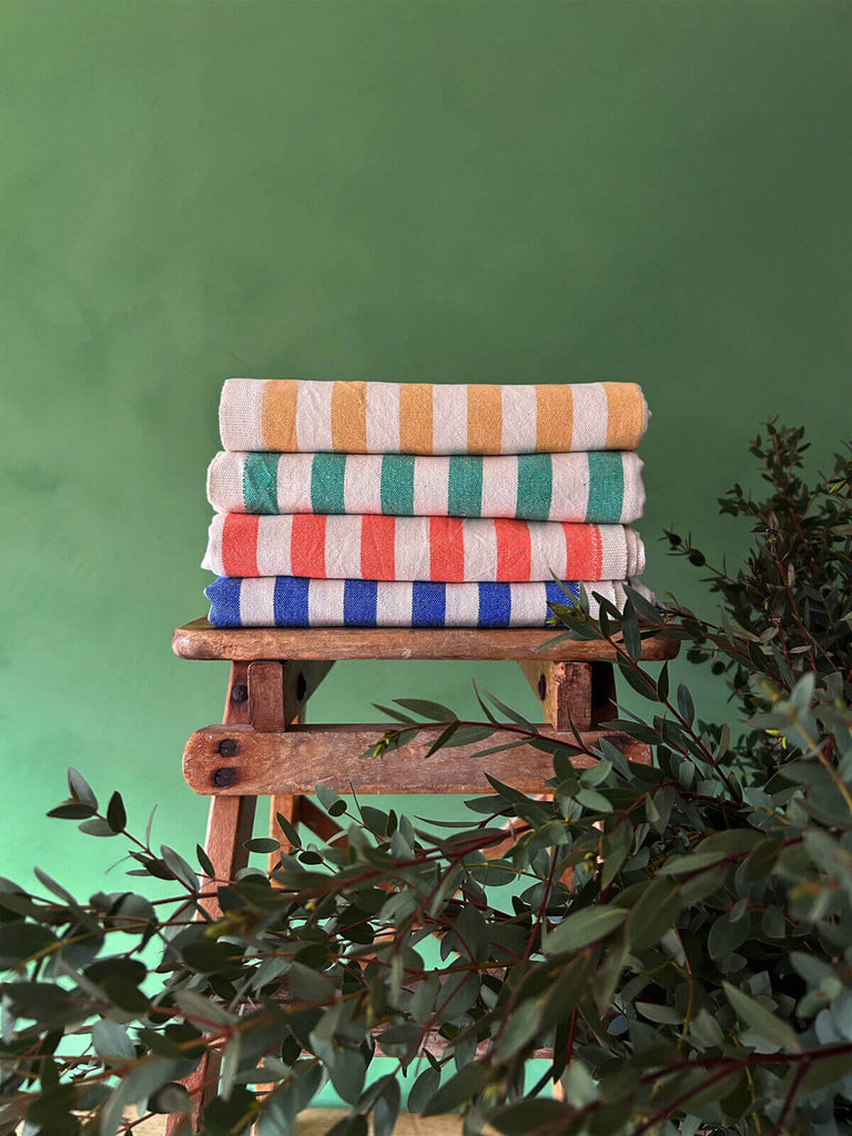 Colourful stack of cotton Brighton Stripe Hammam Towels with bold stripes, against lush greenery by Bihemai Design