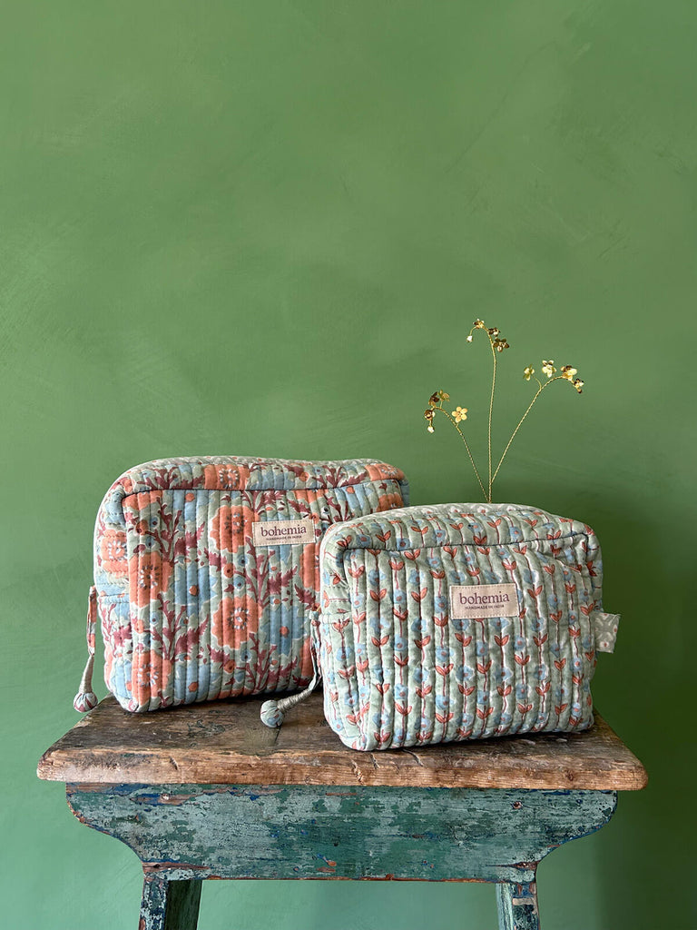 A couple of hand block printed washbags in soft duck egg with blue and terracotta flowers, set against a green textured wall on a rustic wooden stool by Bohemia