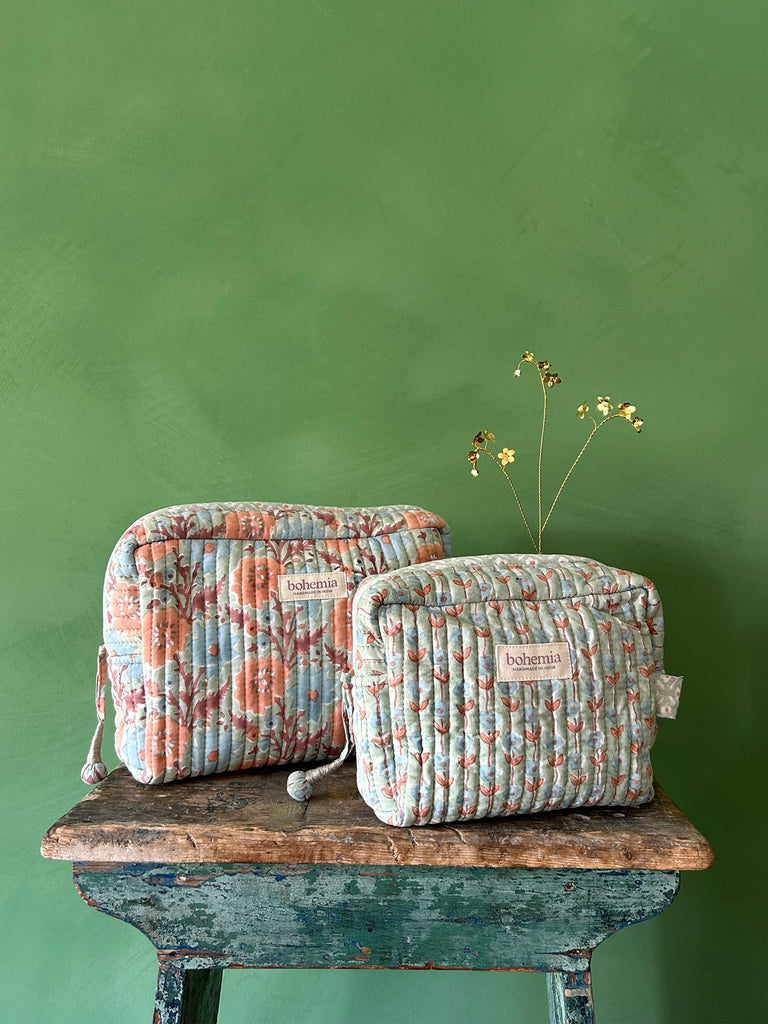 A couple of hand block printed washbags in soft duck egg with blue and terracotta flowers, set against a green textured wall on a rustic wooden stool by Bohemia