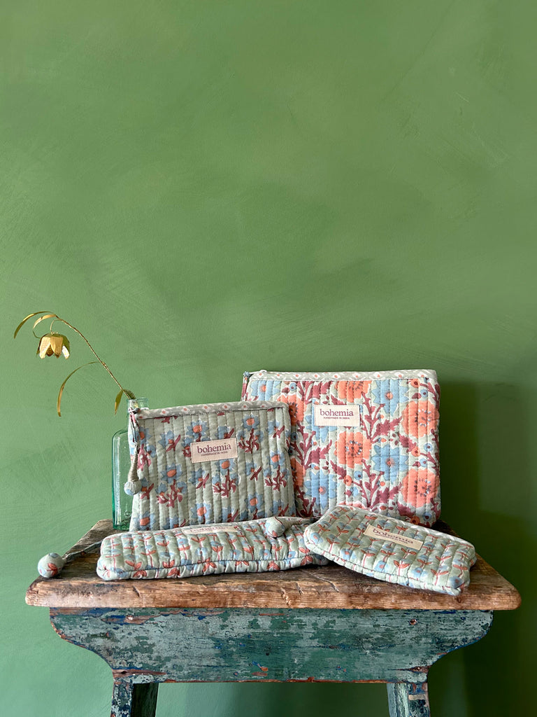 A set of 4 hand block printed zip pouches in delicate duck egg with blue and terracotta flowers, set against a green textured wall on a rustic wooden stool by Bohemia