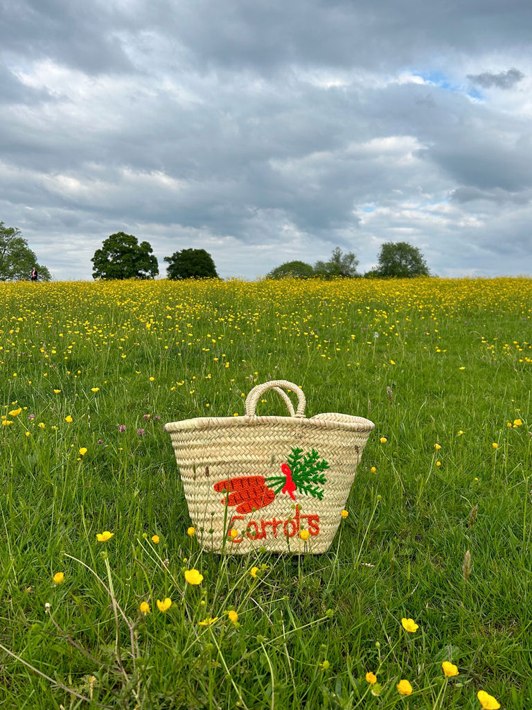 Hand-embroidered carrot basket bag on a green meadow of yellow flowers by Bohemia Design