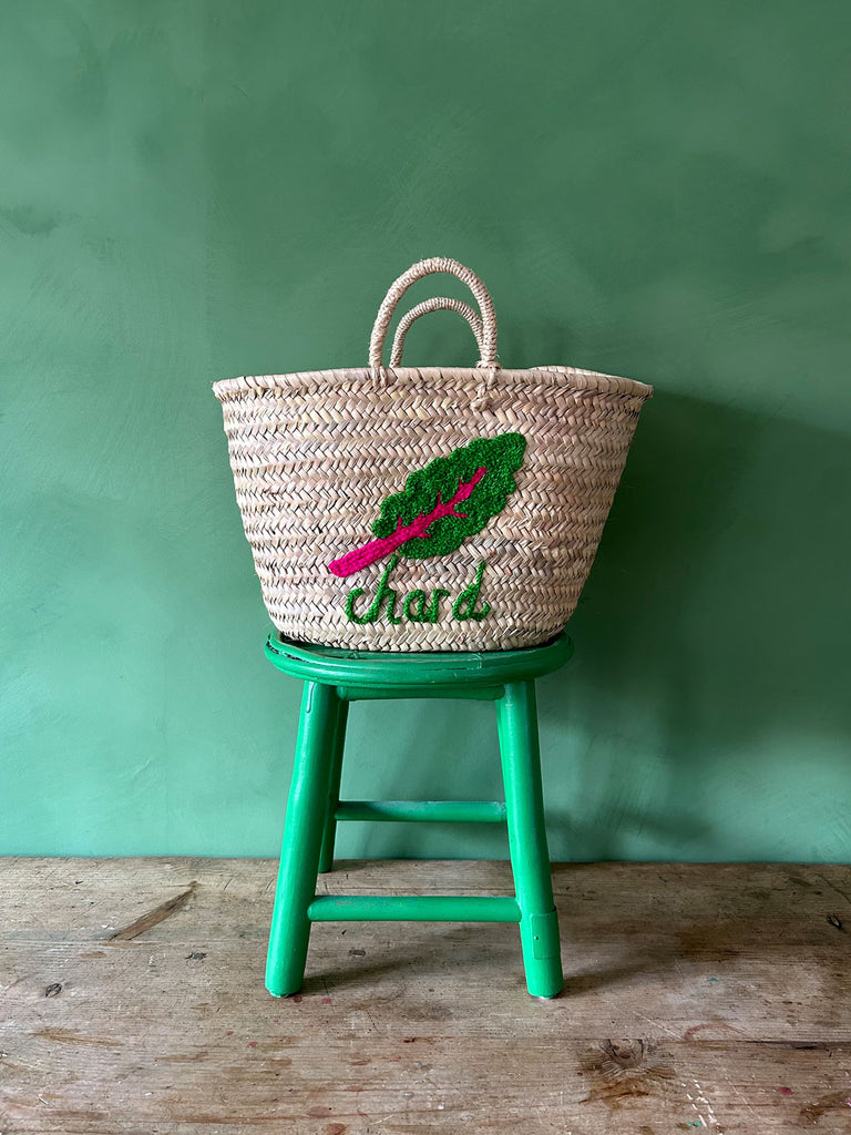 Wholesale handwoven basket bag with an embroidered chard illustration and typography, resting on a green stool against a green wall | Bohemia Design