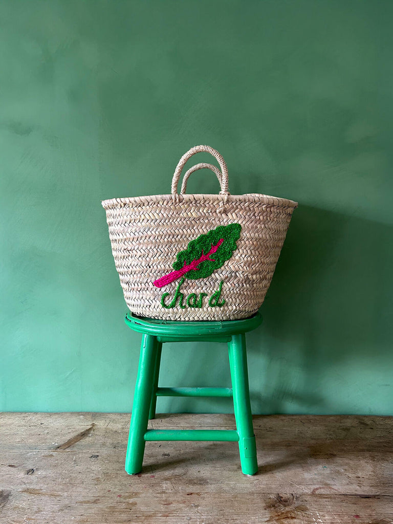 Wholesale handwoven basket bag with an embroidered chard illustration and typography, resting on a green stool against a green wall | Bohemia Design