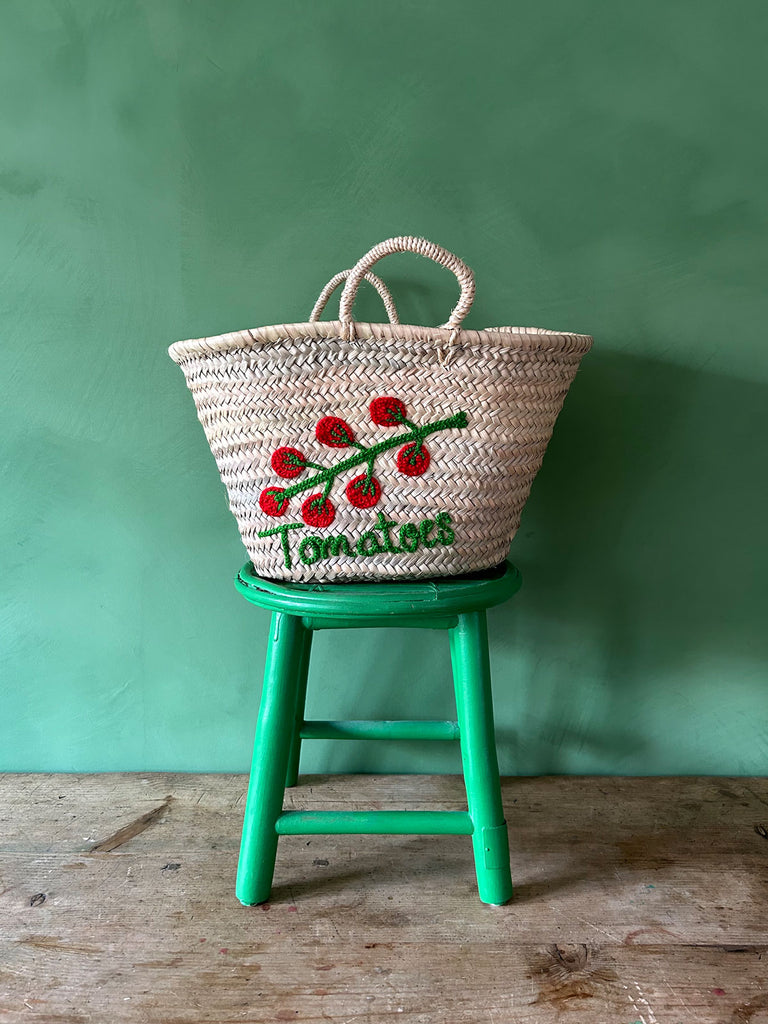 Handwoven wholesale market basket with small handles and an embroidered tomato illustration and accompanying text sitting on a green stool against a green wall | BohemiaDesign