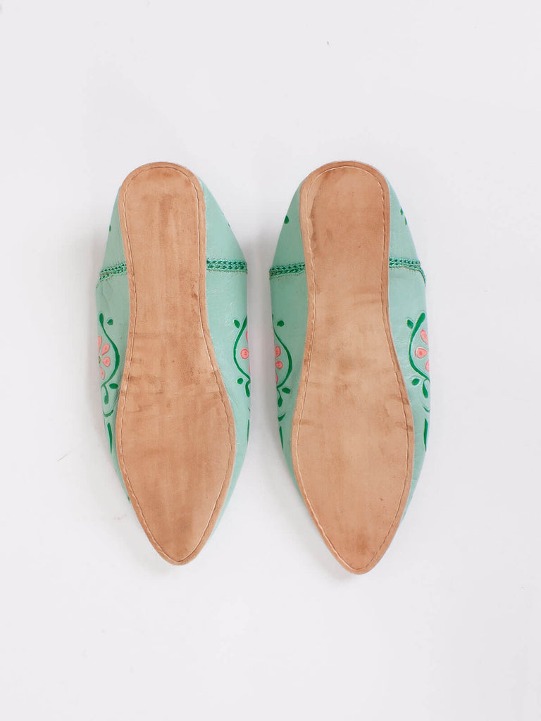 The firm soles of a pair of pointed babouche slippers in a sage green leather with colourful hand painted blooms in pink and green