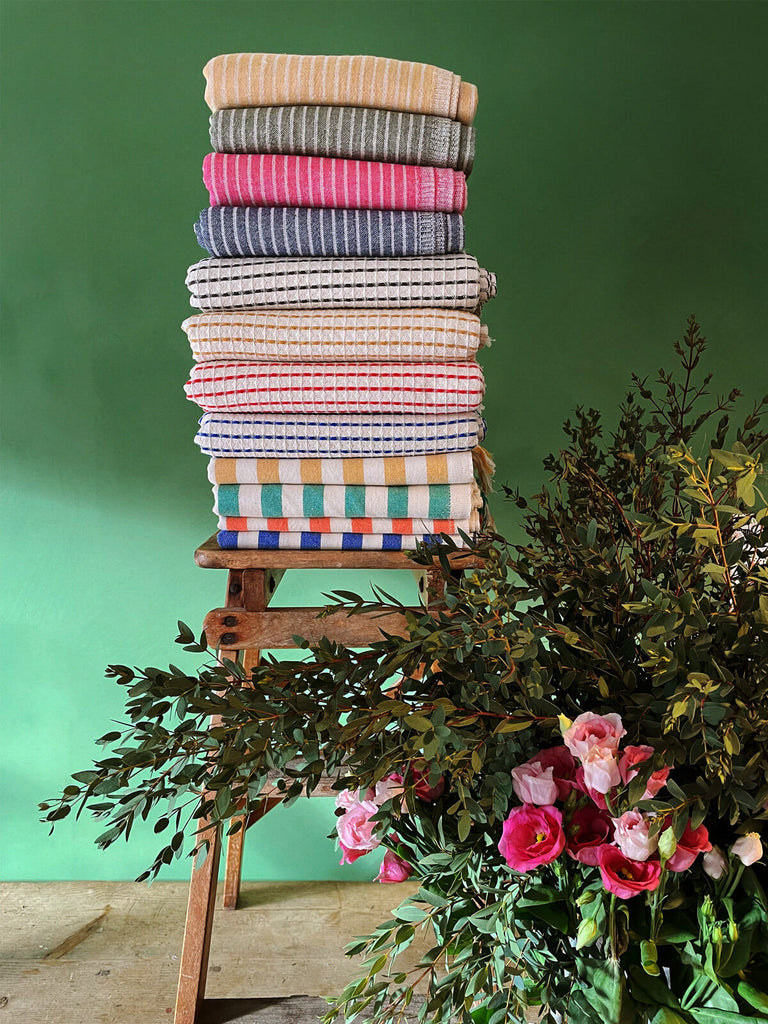 A display of Turkish hammam towels featuring a range of colours, patterns, and textures, neatly folded on a rustic wooden stool | Bohemia Design