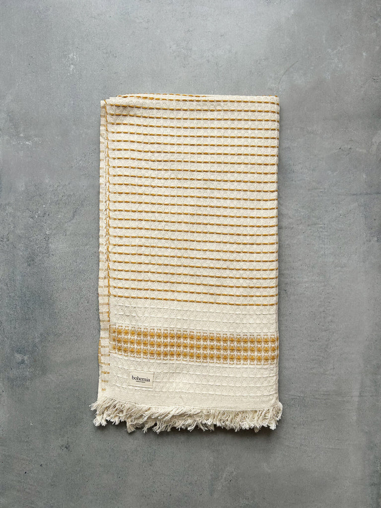 Milos Hammam Towel with a waffle textured weave, featuring a subtle mustard yellow striped pattern and finished with hand-tied tassel fringe on a grey textured background | Bohemia Design