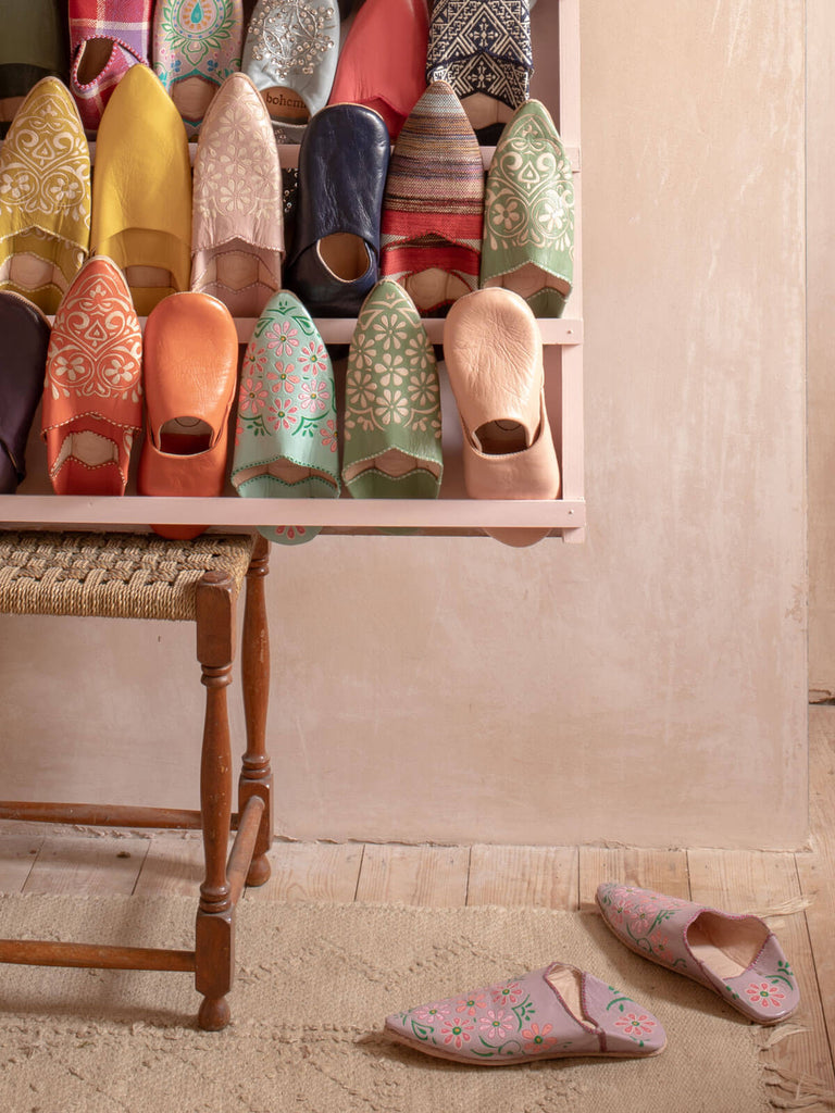 A display of different styles and colours of Bohemia Moroccan babouche slippers in front of a rustic pink plaster wall.