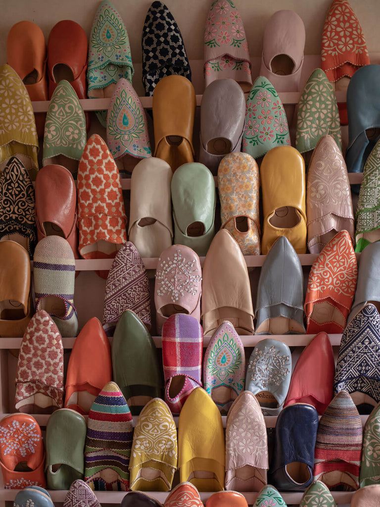 Rows of different styles and colours of Bohemia Moroccan babouche slippers in front of a rustic pink plaster wall.