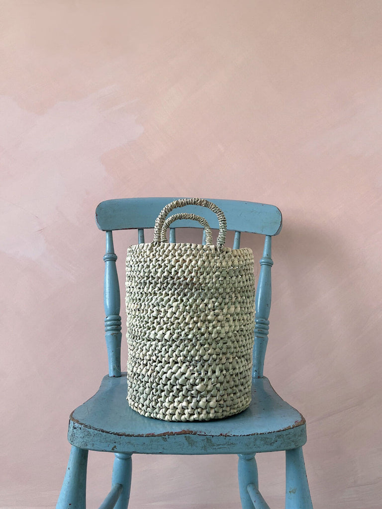 A round basket with short sturdy handles on a blue chair against a plaster pink wall