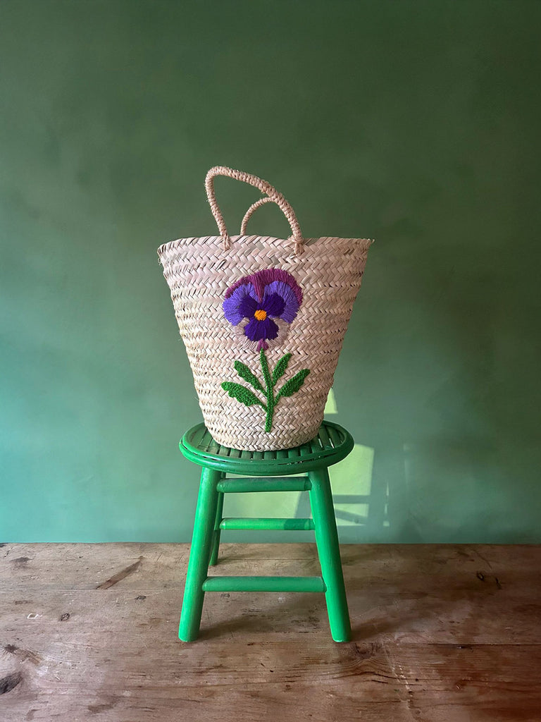 A natural woven basket bag with a hand-embroidered pansy flower displayed on a green stool against a green wall | Bohemia Design Wholesale