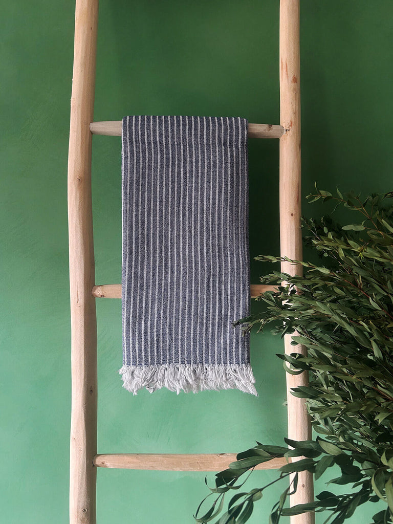 Indigo-blue Portobello hammam towel with subtle white stripes, presented on a rustic wooden ladder against a vibrant green wall | Bohemia Design