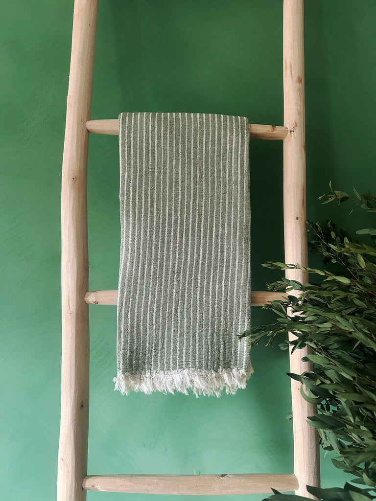 Soft cotton Portobello hammam towel in olive green stripes, presented on a rustic wooden ladder against a vibrant green wall | Bohemia Design