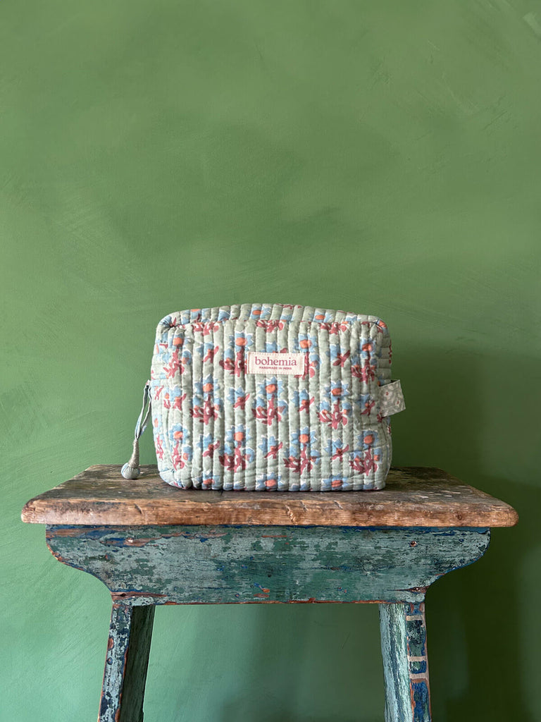 Posie, a cotton quilted washbag with a floral block print in duck egg green, blue, and terracotta, resting on a rustic stool against a green wall