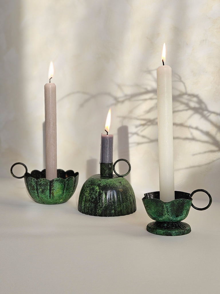 A group of iron candle holders with a vintage moss patina finish, paired with lit candles casting soft shadows on a slightly textured background | Bohemia