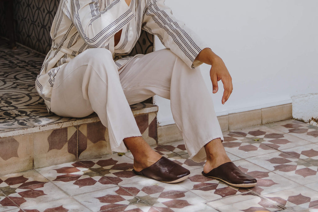 Moroccan babouche leather slippers in chocolate worn by a seated model against Moroccan tiles
