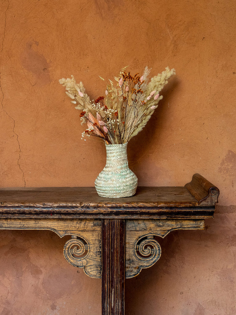 Bohemia Design Palm Leaf Basket Vase with dried flowers on a mantlepiece against a terracotta wall
