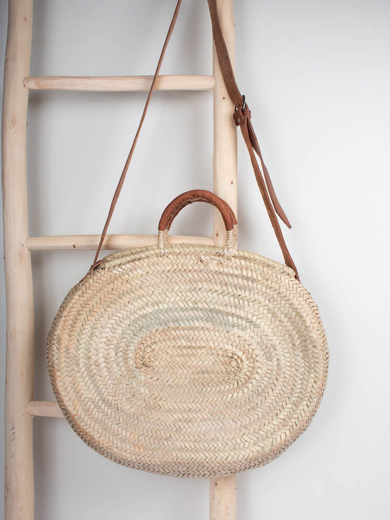 Moroccan woven basket in an oval shape with tan leather strap and handles hanging from a wooden ladder