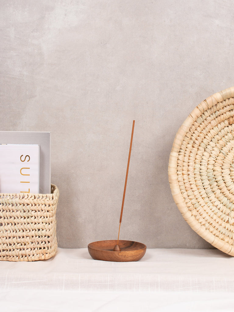 Walnut wood round incense holder by Bohemia Design against a grey wall with palm leaf baskets